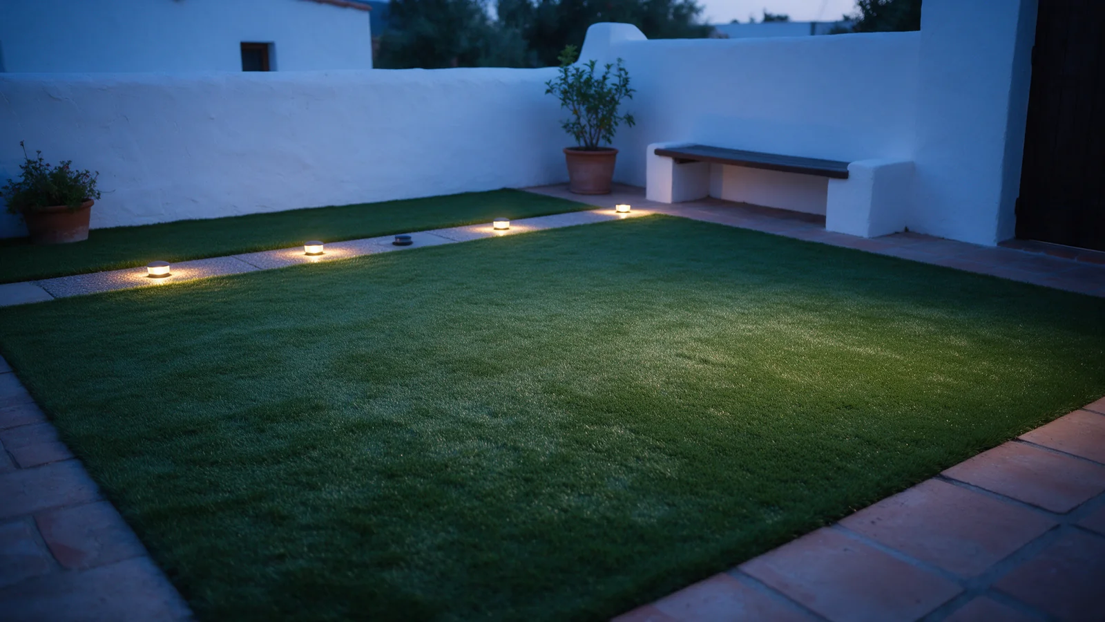 Patio con césped artificial y pequeñas luces cálidas sobre el pavimento al anochecer.