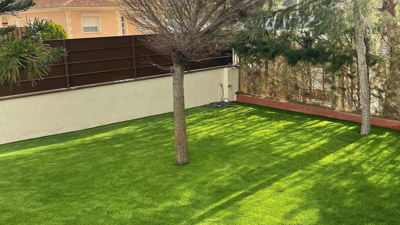 Césped artificial en jardín, árbol y sombras junto a muro